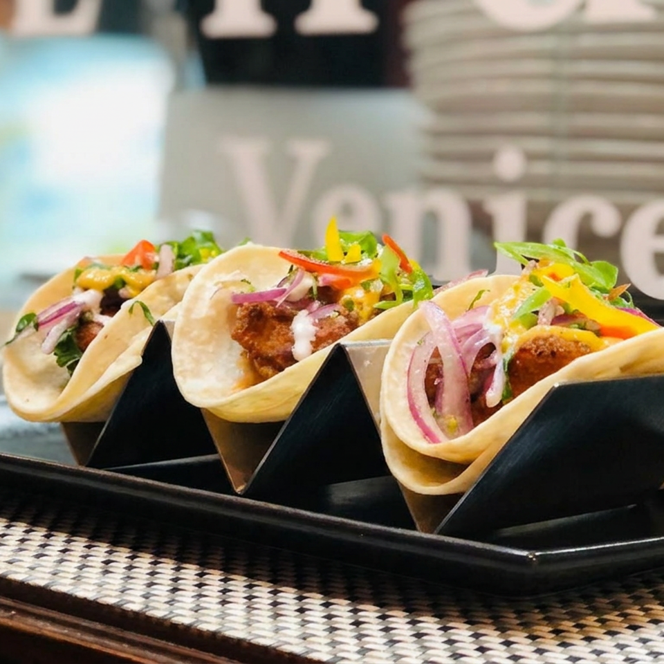 Three colorful tacos with crispy fillings, vegetables, pickled onions, and sauce arranged on a black rectangular plate.