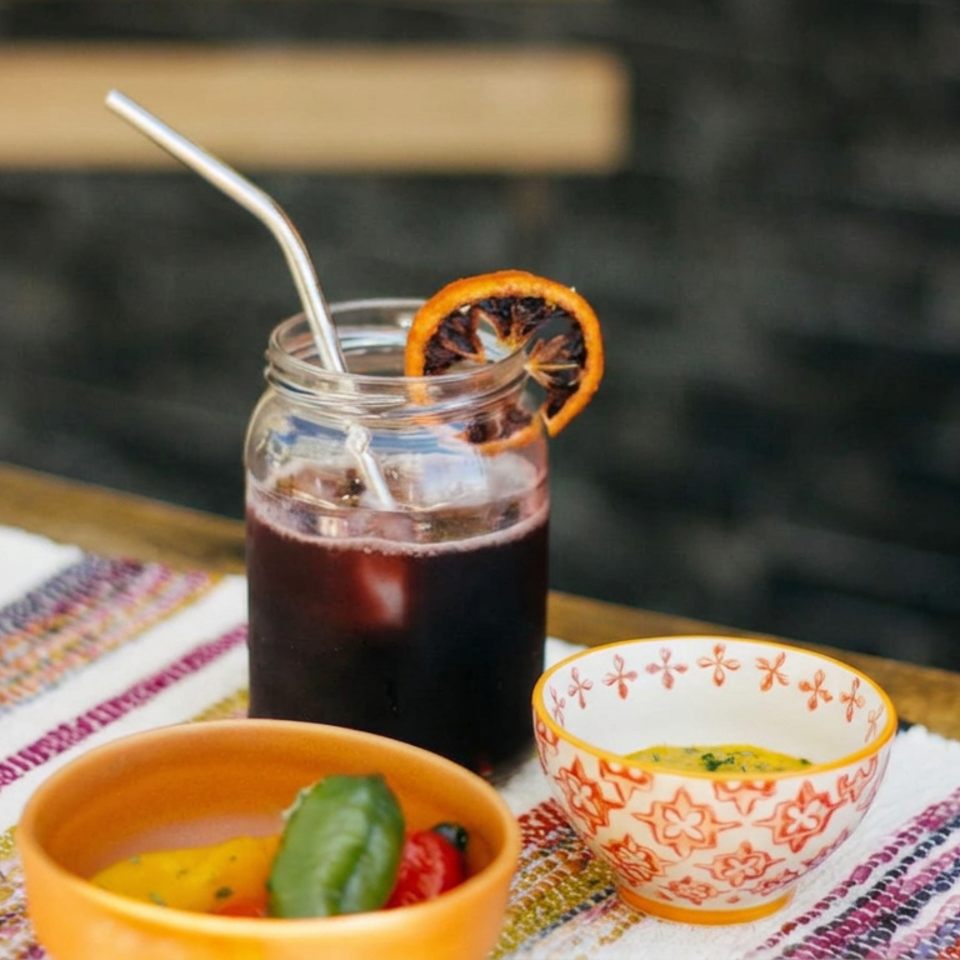 Dark iced beverage in a mason jar with a metal straw and dried orange garnish, alongside colorful bowls on a wooden table.